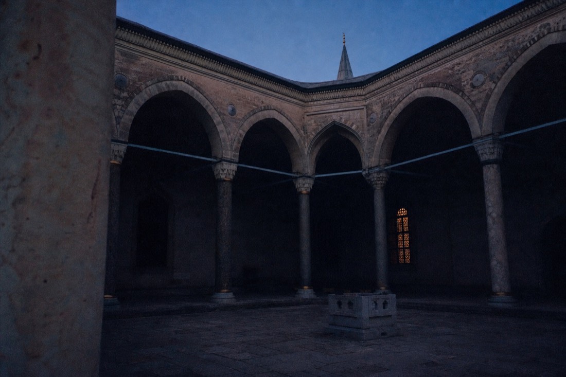 A shadowed arcaded courtyard corner of Topkapi Palace with one lit window at dusk
